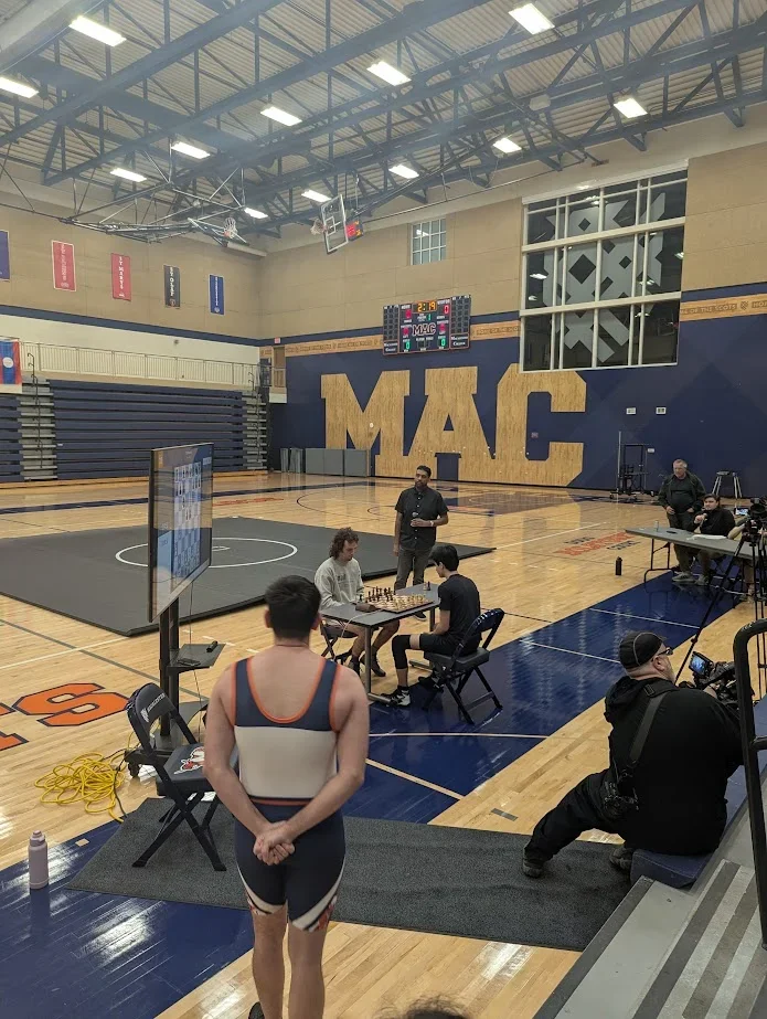 A wrestler looks on as two chesswrestlers play chess on a DGT board in front of a wrestling mat
